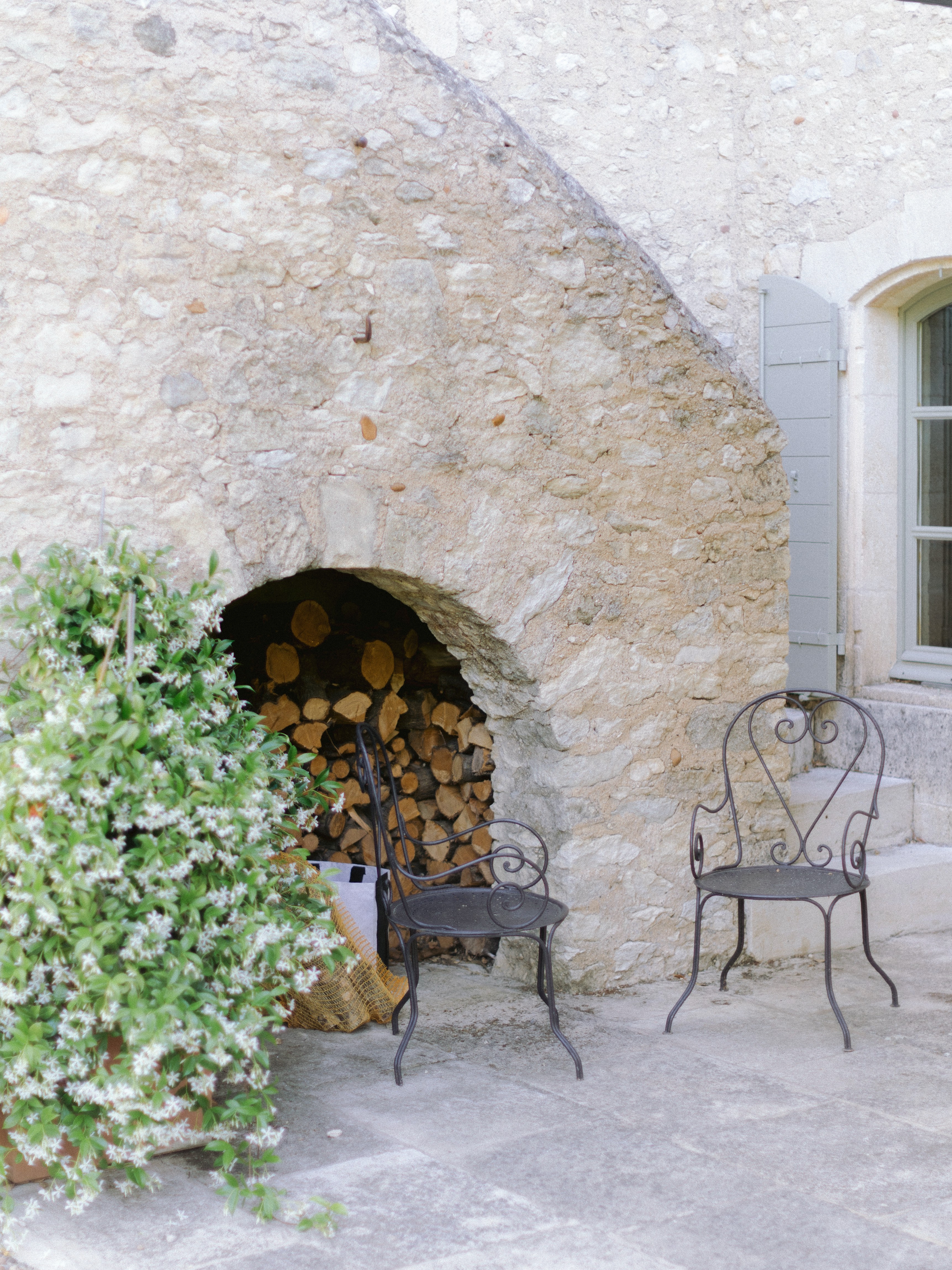 A quaint corner with two metal chairs beside a stone wall featuring an arched alcove filled with firewood, next to blooming white flowers.